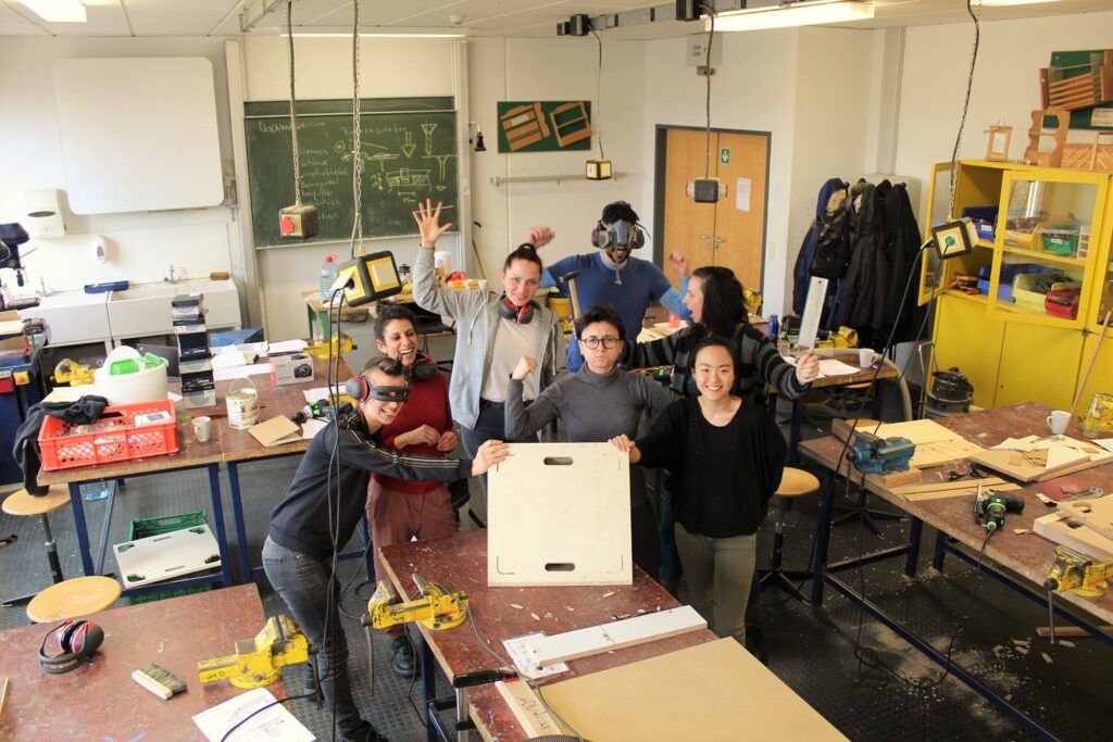 Aus der Vogelperspektive sind 7 junge Menschen fotografiert. Sie stehen zusammen mitten in einer Werkstatt und schauen fröhlich in die Kamera. Sie halten ein Werkstück aus Holz (quadratisch) in den Händen. Eine Person hält einen Hammer und eine anderePaerson hat eine Schweißermaske auf.