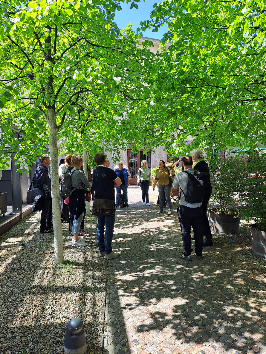 Eine Gruppe von Menschen steht unter Bäumen im Biergarten des Pfefferbergs und schaut interessiert einer Frau zu, die etwas über die in eine Richtung zeigt und etwas dazu erzählt