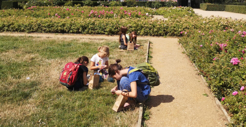Im sommerlichen Garten vor einem Wohngebiet untersuchen Kinder mit und ohne Rucksack den Rasen