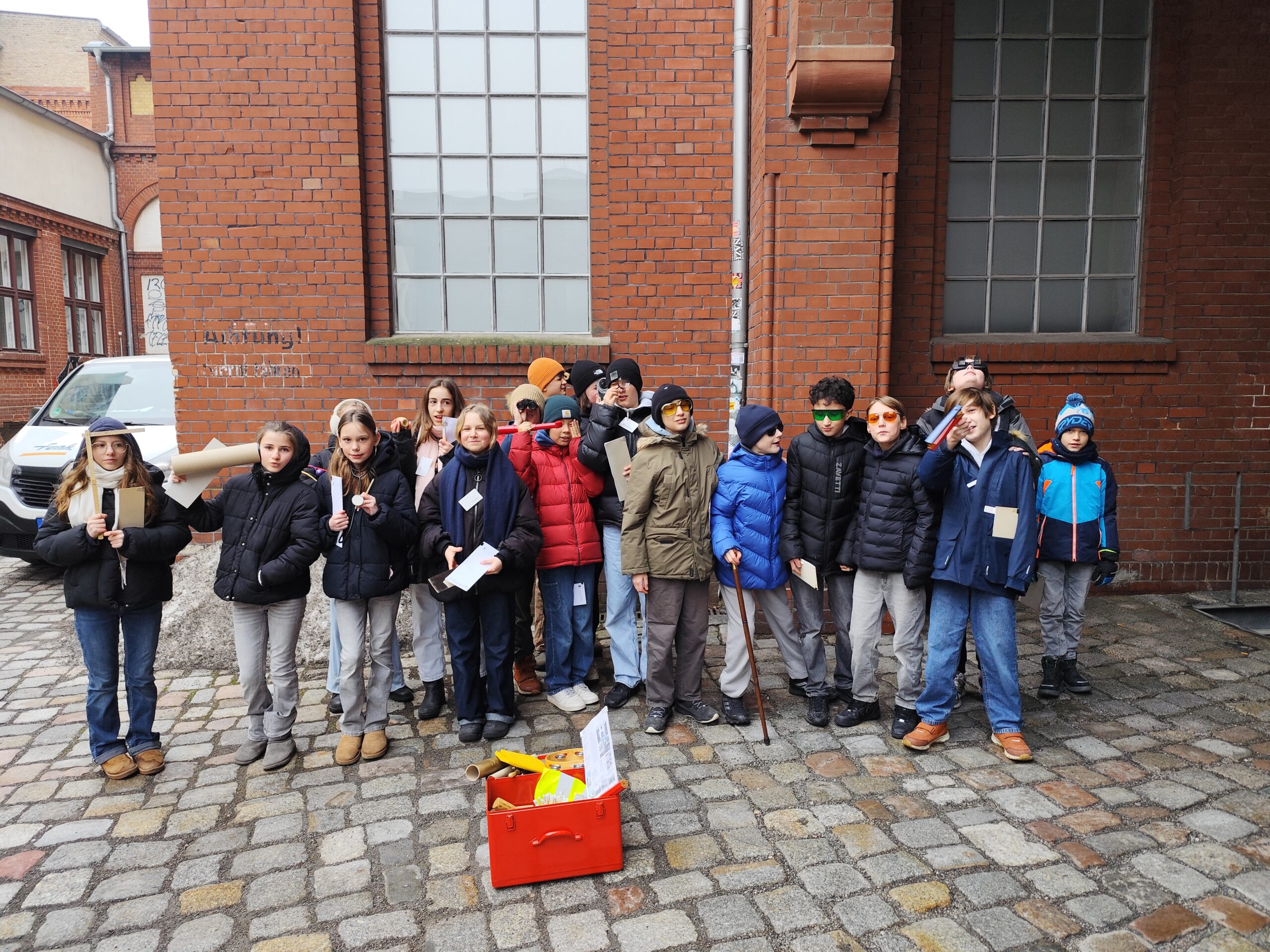 winterlich angezogene Kinder einer Grundschulklasse stehen vor einem roten Backsteingebäude mit roter Klinkerfassade und halten verschiedene Gegenstände in der Hand