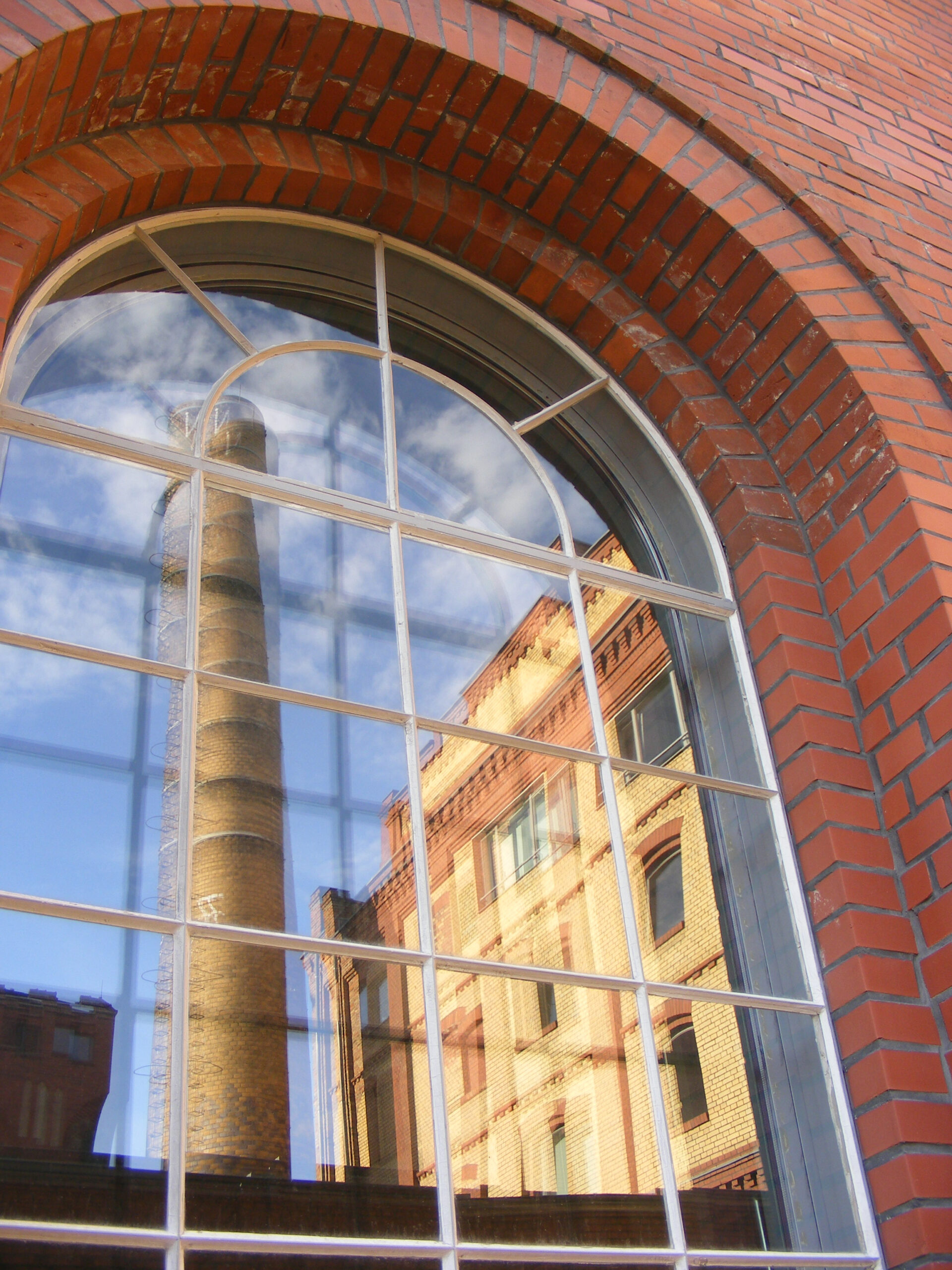 Im Rundfenster einer roten Backstein-Fassade spiegelt sich ein Fabrikschornstein und ein weiteres Gebäude mit gelben und roten Backsteinen