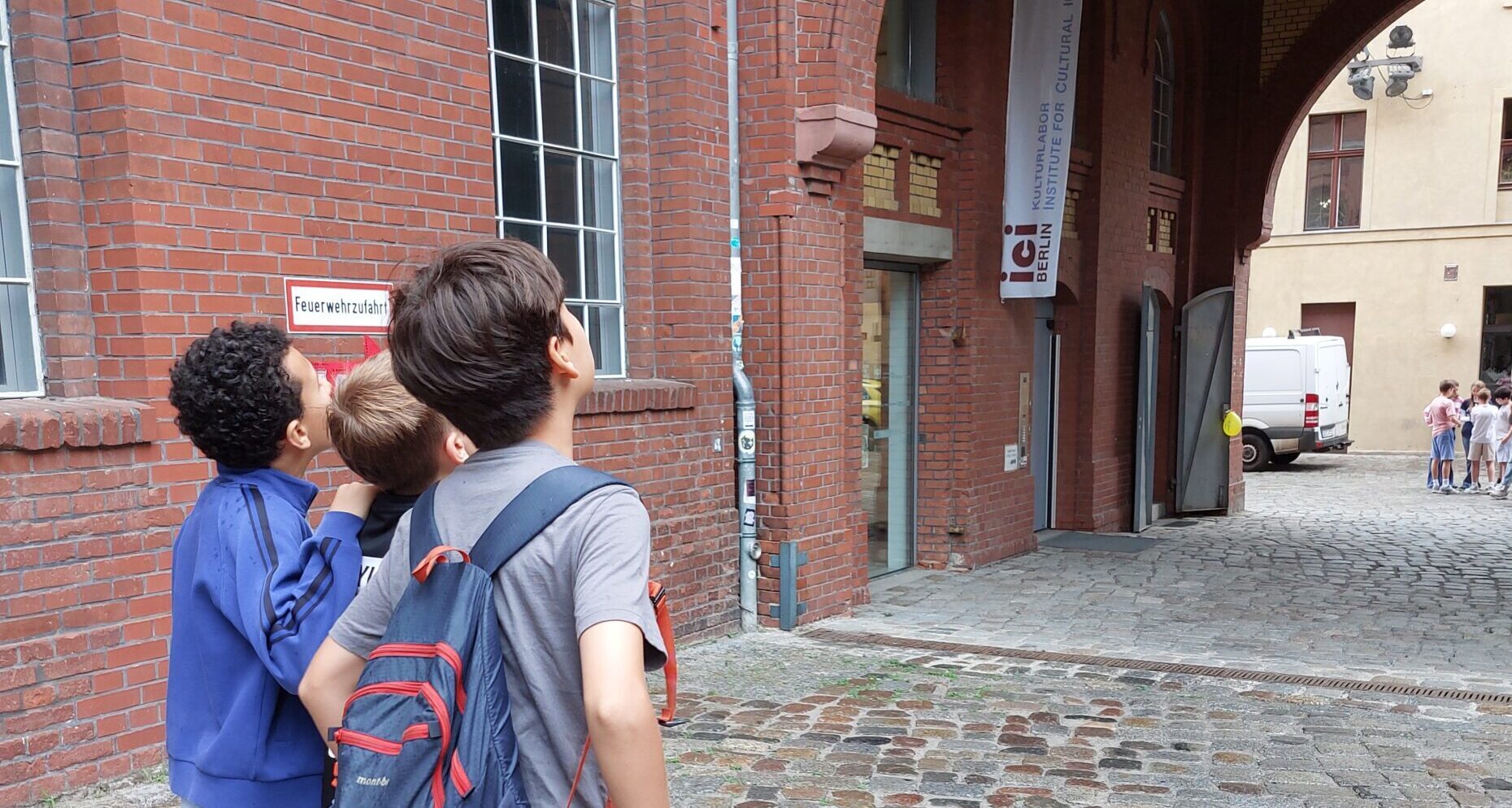 Kinder schauen auf einem historischen gelände mit roter Klinkerfassade nach oben, im Hintergrund steht eine andere Kindergruppe