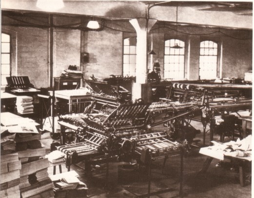 Das historische, bräunliche Foto zeigt die großen, mechanischen Produktionsanlagen der Druckerei "Neues Deutschland". Auch hohe Papierstapel sind zu sehen.