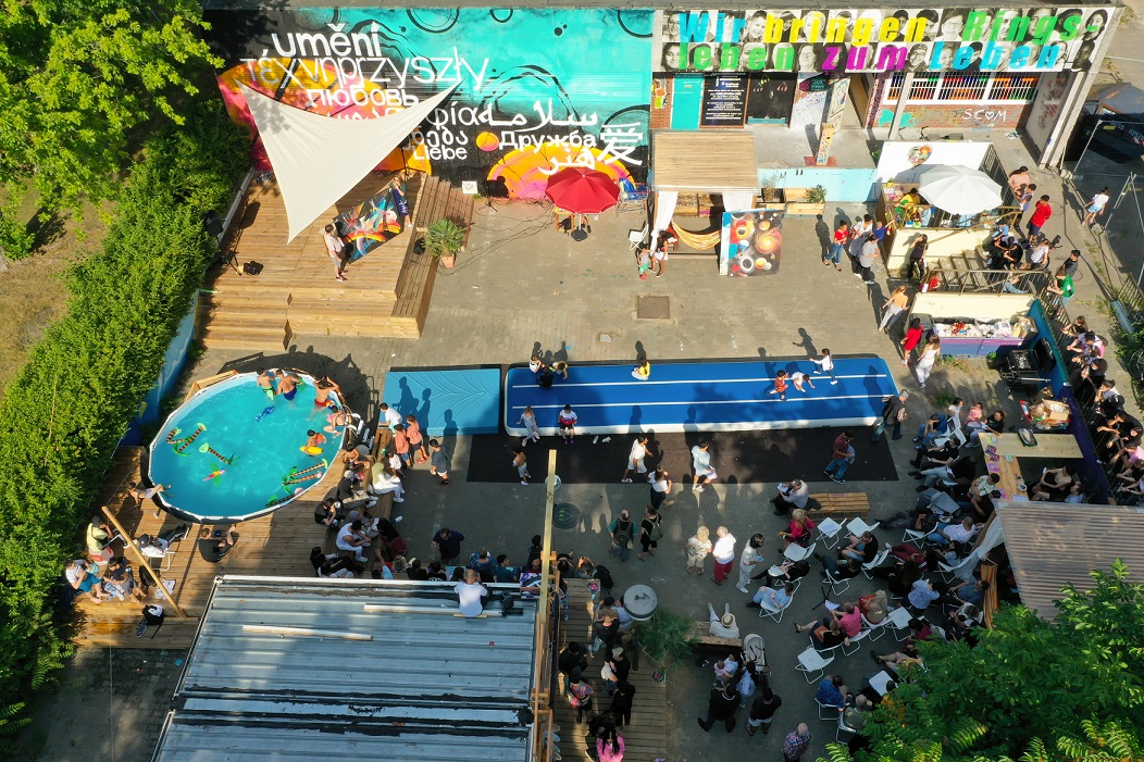Aus der Vogelperspektive sehen wir hinunter auf einen Platz, auf dem in sommerlicher Atmosphäre viele Menschen, jung und alt zusammensetzen, im Pool baden, spielen und Musik hören