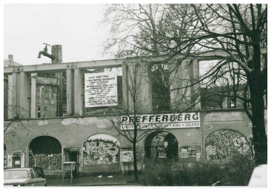 Der Eingang zum Pfefferberg an der Schönhauser Alle 176 in schwarzweiß fotografiert. Man sieht ein Veranstaltungsplakat und mit Graffitti besprühte Rundbögen. Am Schonstein des Gebäudes hinter den Arkaden des Pfefferbergs ist ein riesiger Zapfhahn (Skulptur) zu sehen