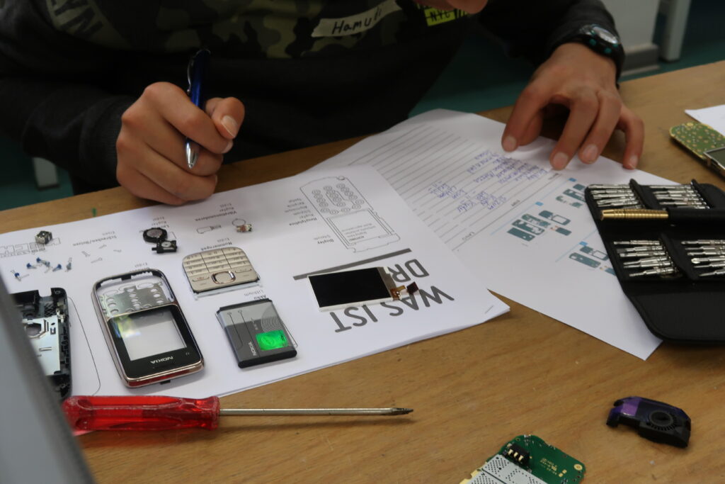 Ein Schüler sitzt an einem Tisch, wo vor ihm die Teile eines auseinandergebauten Handys ausgebreitet liegen, geordnet auf einem Bauteile-Plan. Auf einem Papier-Formular listet er genau sein Vorgehen auf. Auf dem Arbeitstisch sieht man seine mechanischen Feinwerkzeuge.