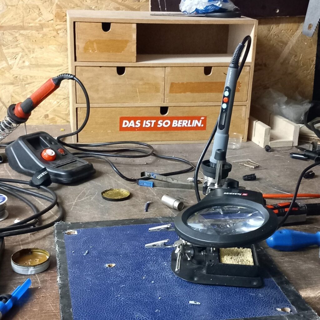 Auf einer Arbeitsstation an einer Schule für Löt-Arbeiten befinden sich zwei Lötgeräte, eine Lupe und sonstige Gegenstände wie Kabel, Schmierfett und Schraubendreher